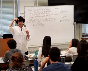 Hildy Gottlieb stands next to a white board, teaching a workshop. There are community members in desks facing her.