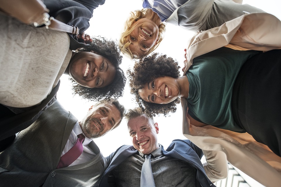 5 people in business attire, standing in a circle, looking down into the center of the circle. The camera is on the ground, looking up at their smiling faces.