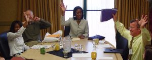 Board table with 2 Black women and 2 white men, all smiling and raising their hands. 