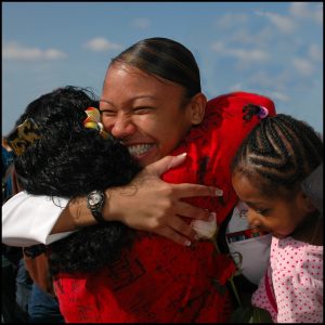 A sailor on leave hugs her children, seeing them for the first time. She has a huge smile on her face.