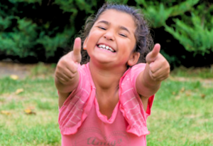 Happy little girl with a pink shirt, pointing at you, grinning so big her teeth are showing
