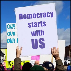 Poster at a protest march, held above the crowd, that says "Democracy starts with US"