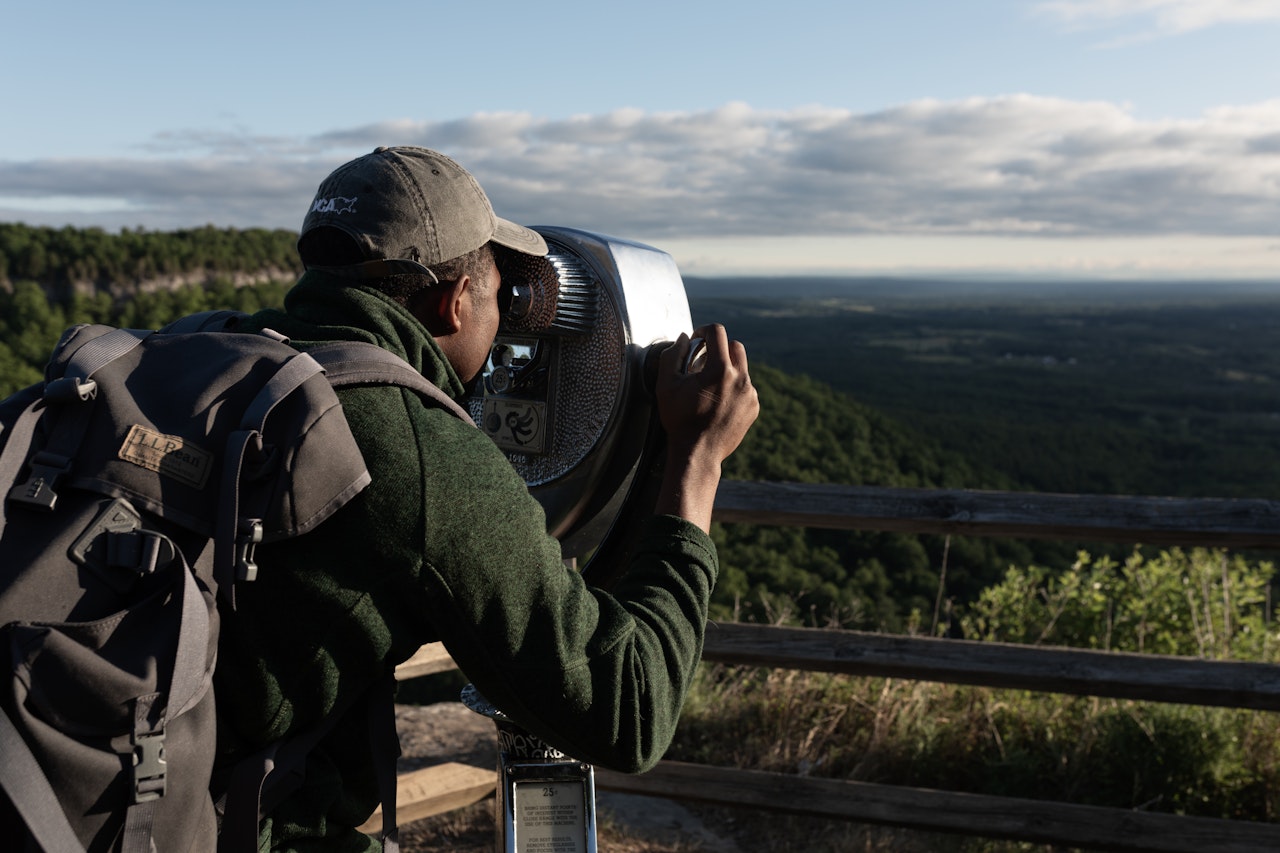 Man with backpack, at a scenic vista point, looking through a telescope at the tree-lined view in the distance
