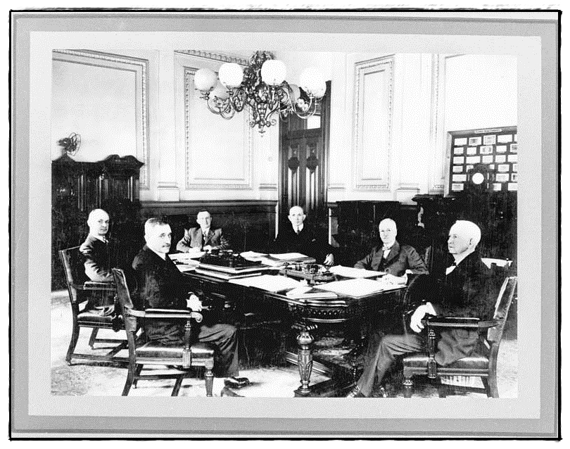 1880's era B&W photo of a group of old white men in formal business attire, sitting around a board table