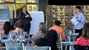 Hildy on the right, May Miller-Dawkins on the left, in front of workshop attendees. They are standing in front of a tan cloth background with a black pattern