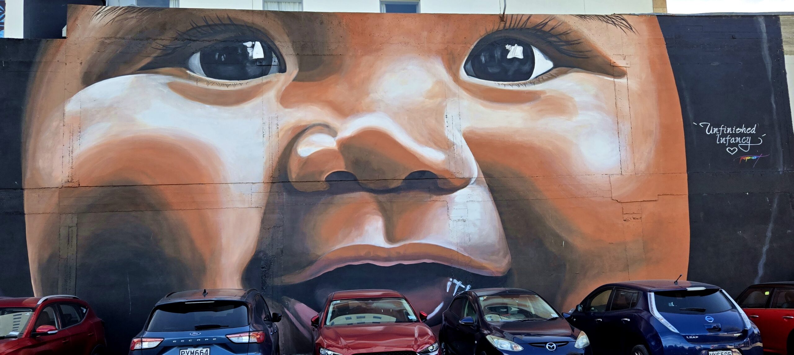 Large mural taking up 6 car-widths in a parking lot, image is a happy baby with golden brown skin and dark brown eyes.