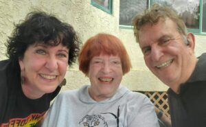 Hildy on the left with curly dark hair and a dark t-shirt, Ann in the middle with straight red hair and a white sweatshirt, Dimitri on the right with curly grey hair and black polo shirt. All are smiling.
