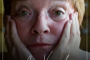 Close-up Photo of Ann Vermel. A white woman of a certain age, red hair, pink cheeks, holding her head in her hands. The text overlay says, "Ann Vermel, in loving memory."