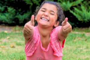 Happy little girl with a pink shirt, pointing at you, grinning so big her teeth are showing