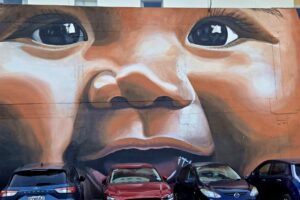 Large mural taking up 6 car-widths in a parking lot, image is a happy baby with golden brown skin and dark brown eyes.